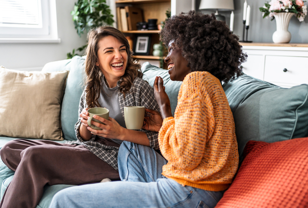 Two female friends laughing and drinking coffee on sofa at home.